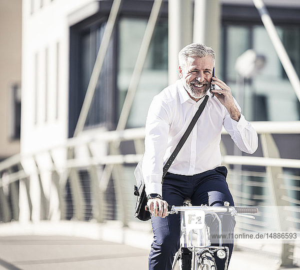 Glücklicher reifer Geschäftsmann  der auf einer Brücke in der Stadt mit dem Handy telefoniert und Fahrrad fährt