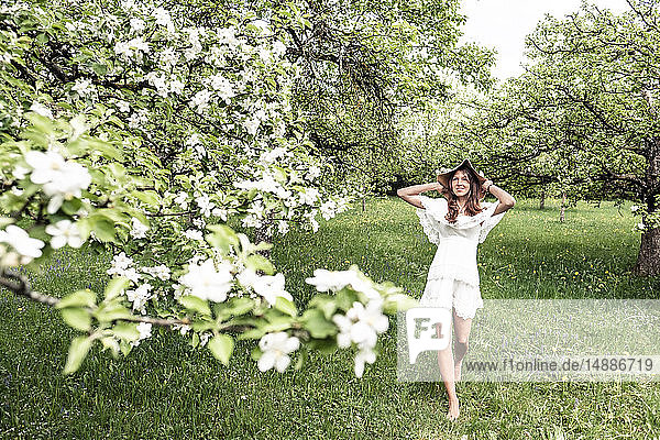 Junge Frau in weißem Kleid und Schlapphut geht barfuß im Garten mit blühenden Apfelbäumen