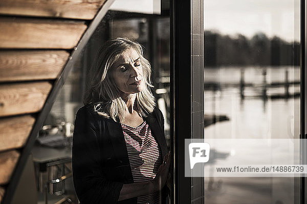 Geschäftsfrau  die auf einem Hausboot steht und mit geschlossenen Augen die Sonne am Fenster genießt