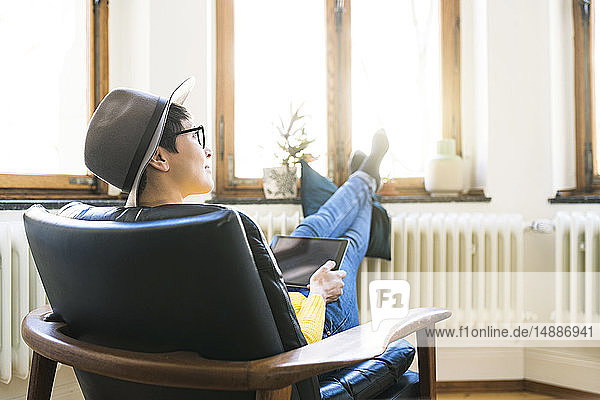 Woman relaxing in lounge chair holding a tablet and looking outside windows in stylish apartment
