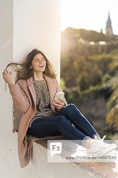 Lachende junge Frau sitzt auf einer Wand und hält ein Handy