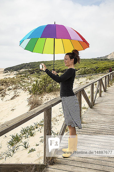 Frau mit buntem Regenschirm steht am Strand und nimmt einen Selfie