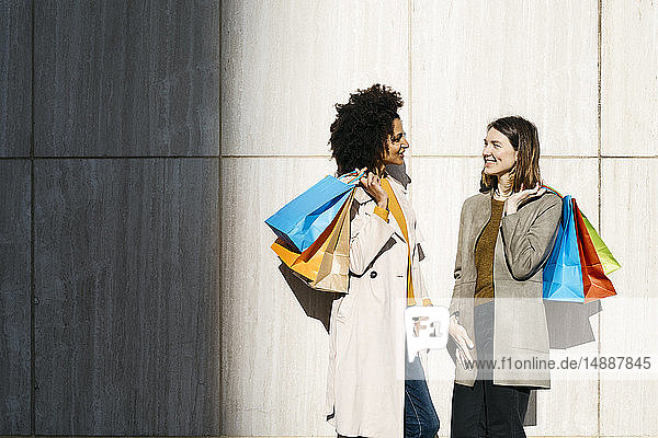 Zwei glückliche Frauen mit Einkaufstaschen stehen an einer Wand und unterhalten sich