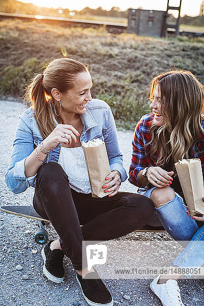 Zwei lachende Freunde sitzen auf dem Longboard und essen Popcorn
