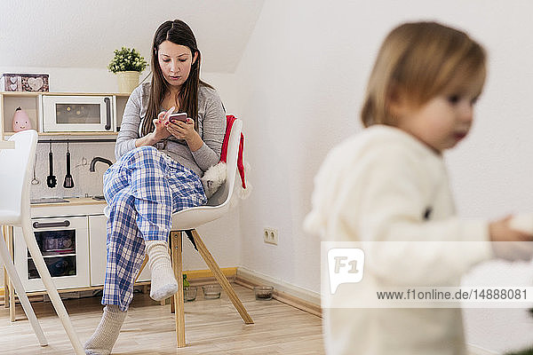 Frau sitzt in der Küche und benutzt ein Smartphone  während die kleine Tochter im Vordergrund steht