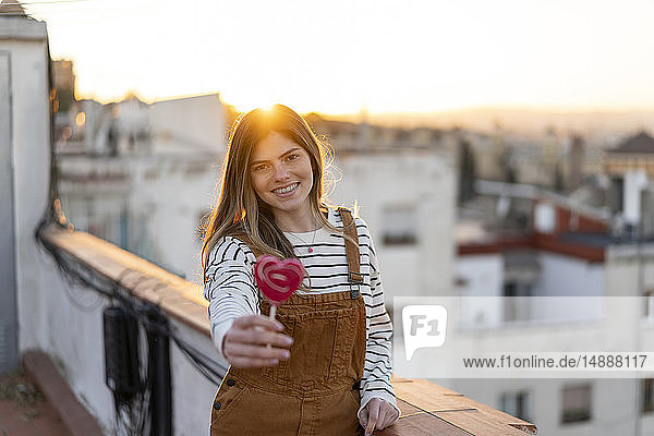 Porträt einer lächelnden jungen Frau  die bei Sonnenuntergang einen roten Lutscher auf der Dachterrasse schenkt