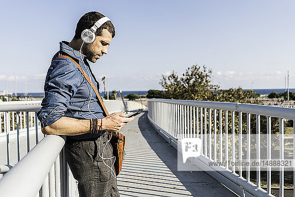 Mann steht auf Steg und hört Musik mit Kopfhörern und Smartphone