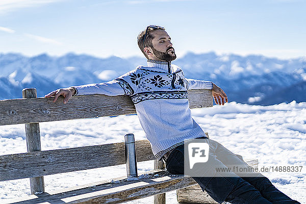 Deutschland  Bayern  Brauneck  Mann sitzt im Winter auf einer Bank in den Bergen und macht Pause