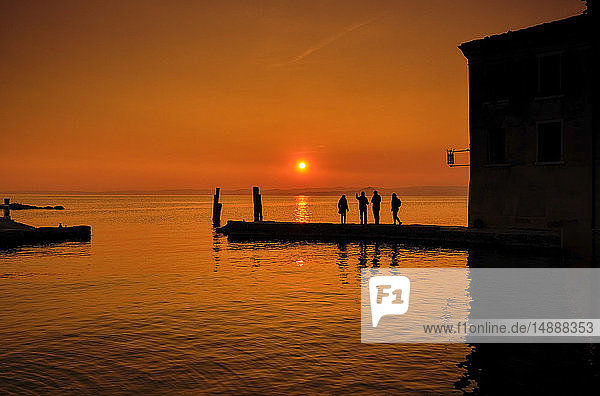 Italien  Punta san Vigilio  Silhouetten von vier Personen auf einem Steg  die im Winter den Sonnenuntergang über dem Gardasee beobachten