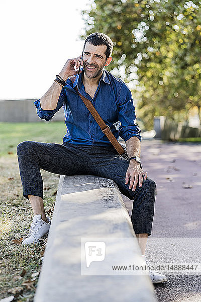 Porträt eines lächelnden Mannes am Telefon  der im Freien an einer Wand sitzt