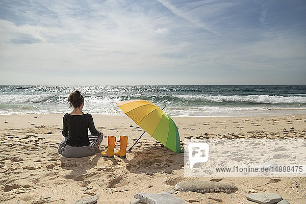 Frau mit buntem Sonnenschirm am Strand sitzend  Rückansicht