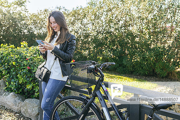 Smiling young woman with bicycle in park using cell phone