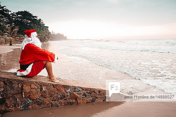 Thailand  als Weihnachtsmann verkleideter Mann sitzt bei Sonnenuntergang auf einer Mauer am Strand