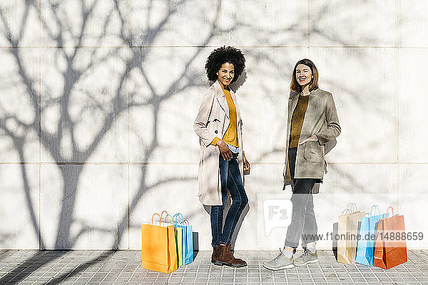 Porträt von zwei glücklichen Frauen mit Einkaufstaschen an einer Wand stehend