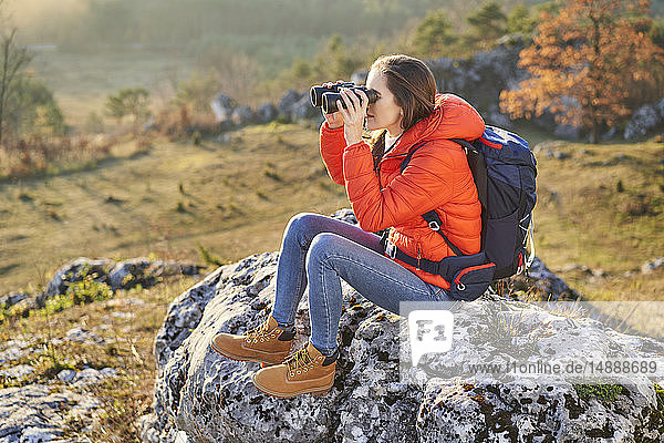 Frau auf einer Wanderung in den Bergen auf einem Felsen sitzend und durch ein Fernglas blickend