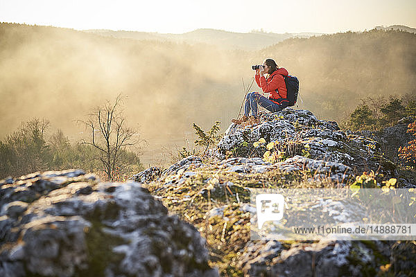 Frau auf einer Wanderung in den Bergen auf einem Felsen sitzend und durch ein Fernglas blickend