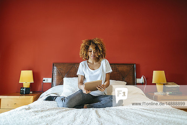Junge Frau mit lockigem Haar sitzt mit Tablette auf dem Bett