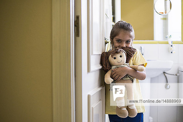 Porträt eines lächelnden kleinen Mädchens  das zu Hause im Türrahmen steht und eine Puppe hält
