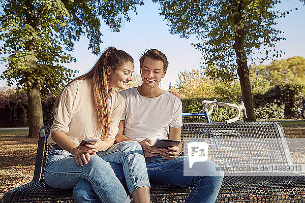Young couple in park sitting on a bench with cell phone and tablet