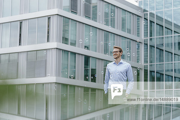 Young businessman standing in front of modern office building  watching soap bubbles