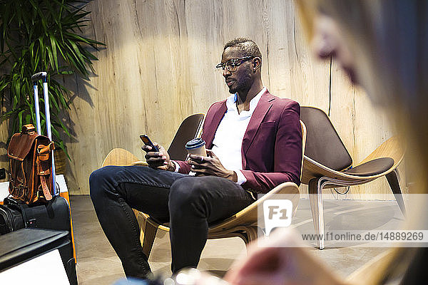 Geschäftsmann mit Gepäck sitzt in der Hotellobby  benutzt ein Smartphone  trinkt Kaffee