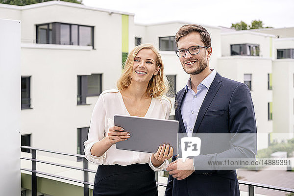 Immobilienmakler steht mit einem Kunden auf einem Balkon und schaut auf ein digitales Tablett