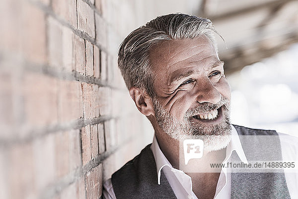 Porträt eines lächelnden  reifen Geschäftsmannes  der an einer Ziegelmauer lehnt und zur Seite schaut