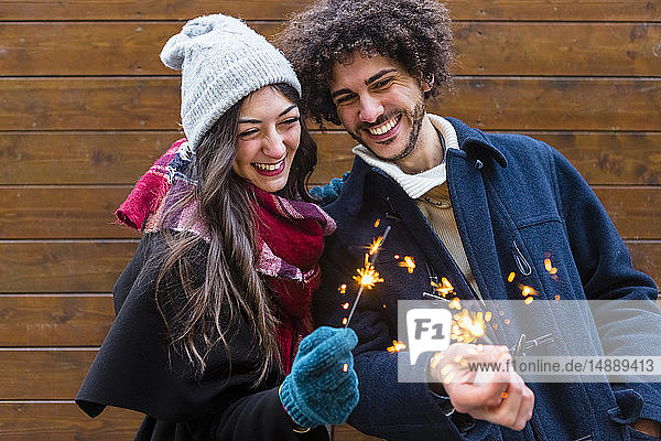 Happy young couple in winterwear holding sparklers in front of wooden wall