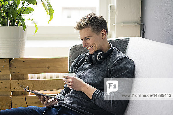 Lächelnder junger Mann mit Tablette und Kopfhörer auf Couch sitzend