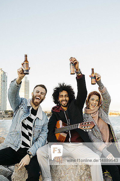 Porträt von drei glücklichen Freunden mit Gitarre  die bei Sonnenuntergang an der Küste Bierflaschen hochheben