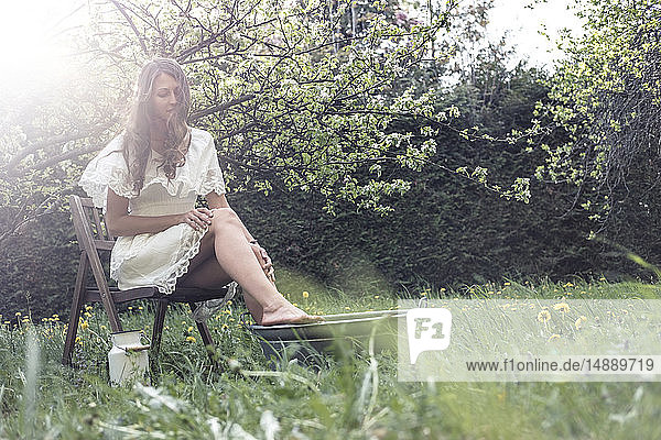 Junge Frau in weißem Kleid sitzt im Garten und nimmt ein Fußbad