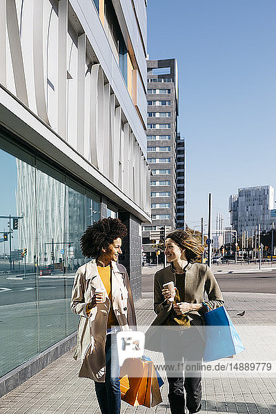 Zwei glückliche Frauen mit Einkaufstaschen und Kaffee zum Mitnehmen spazieren durch die Stadt
