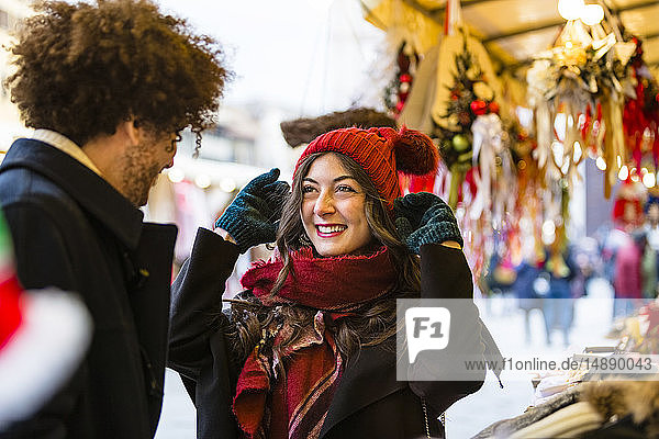 Glückliches junges Paar auf dem Weihnachtsmarkt mit Frau  die einen Wollhut anprobiert