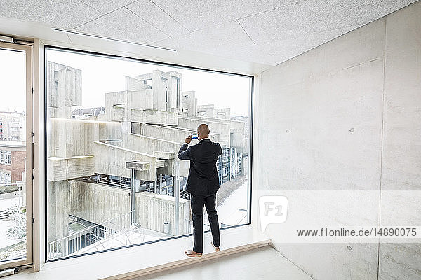 Back view of barefoot businessman standing on window sill taking picture with smartphone
