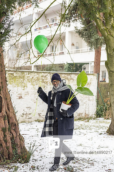 Lächelnder reifer Mann mit grünem Luftballon und Topfpflanze im Winter