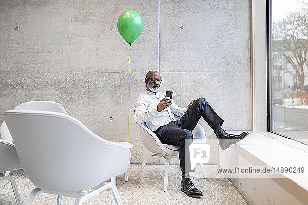 Reifer Geschäftsmann mit grünem Ballon sitzt auf einem Sessel und schaut auf sein Handy