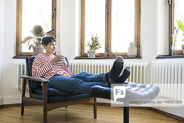 Kurzhaarige Frau entspannt sich in Lounge Chair mit Kaffeetasse in stilvoller Wohnung