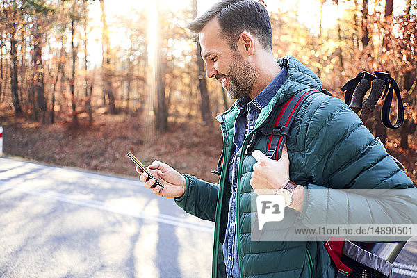Lächelnder Mann überprüft Smartphone auf einer Straße im Wald während einer Rucksacktour