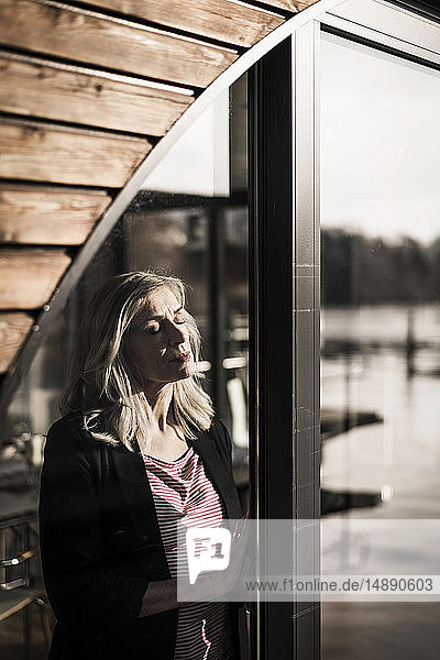Geschäftsfrau  die auf einem Hausboot steht und mit geschlossenen Augen die Sonne am Fenster genießt