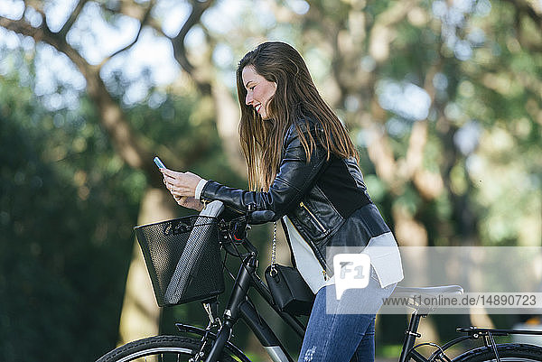 Lächelnde junge Frau mit Fahrrad im Park mit Mobiltelefon