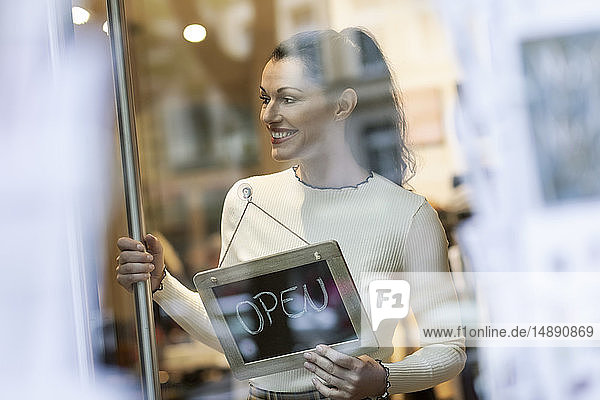 Reife Frau steht im Geschäft  lächelt  mit offenem Schild im Fenster