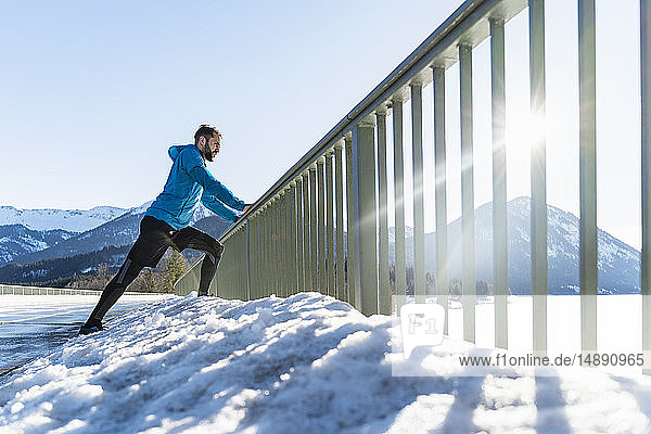 Deutschland  Bayern  sportlicher Mann streckt sich im Winter auf dem Brückengeländer