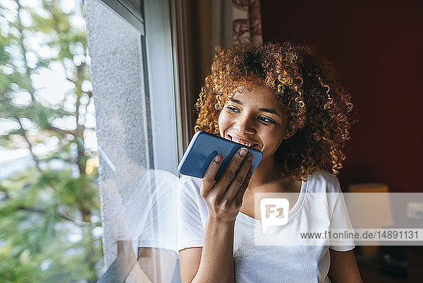 Young woman with curly hair sending voice message with cell phone next to the window