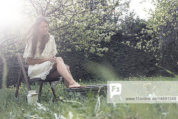 Junge Frau in weißem Kleid sitzt im Garten und nimmt ein Fußbad
