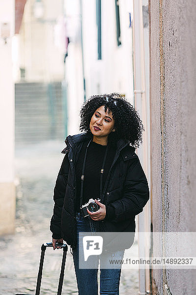 Young woman holding camera and suitcase in alley