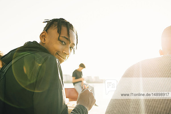 Porträt eines lächelnden Teenagers  der eine Mahlzeit hält  während er mit einem Freund an einem sonnigen Tag auf der Promenade sitzt