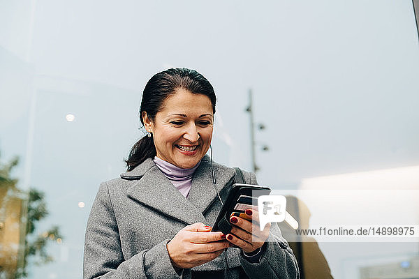Niedriger Blickwinkel auf eine lächelnde Geschäftsfrau  die ein Smartphone benutzt  während sie gegen ein Gebäude in der Stadt steht