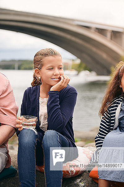 Lächelndes Mädchen schaut weg  während es am Seeufer mit der Familie im Park beim Picknick ein Getränk hält