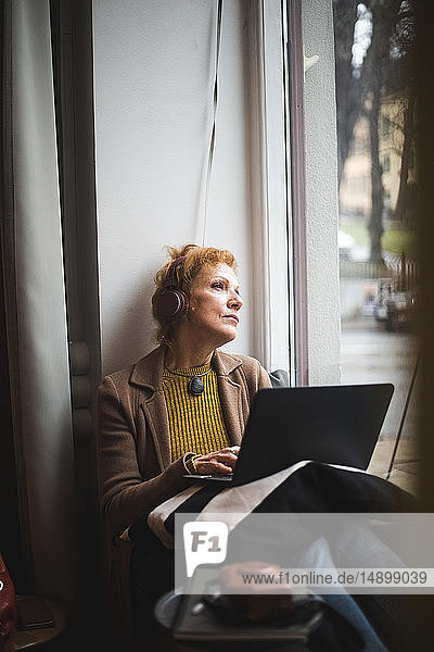 Nachdenkliche reife Berufstätige schaut weg  während sie mit dem Laptop auf dem Fensterbrett im Büro sitzt
