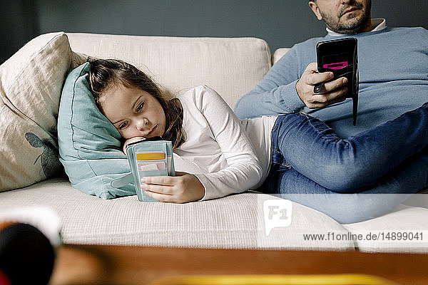 Father and daughter using mobile phones on couch at home